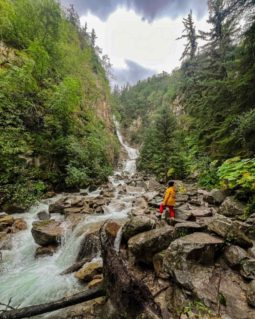 Alina at the base of Lower Reid Falls in Skagway, Alaska. There is a long, narrow waterfall cutting through boulders and trees on either side. Alina wears a black beanie, a yellow rain jacket, and red leggings.