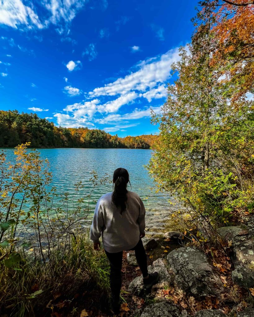 Alina with her back to the camera, overlooks Pink Lake in Gatineau Park. The lake is a bright blue, and the trees have their fall foliage.
