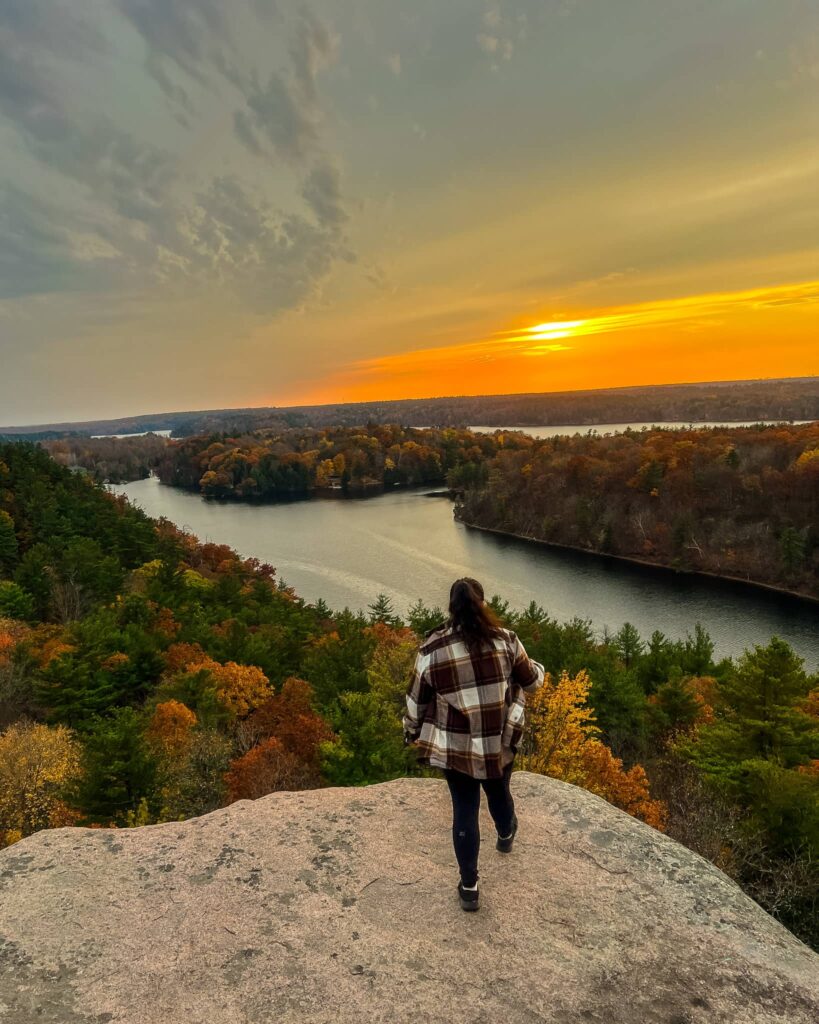 Alina, in a brown plaid shirt, stands with her back to the camera. She is overlooking the bay at Rock Dunder summit. The sun is setting and the trees have their fall colours.