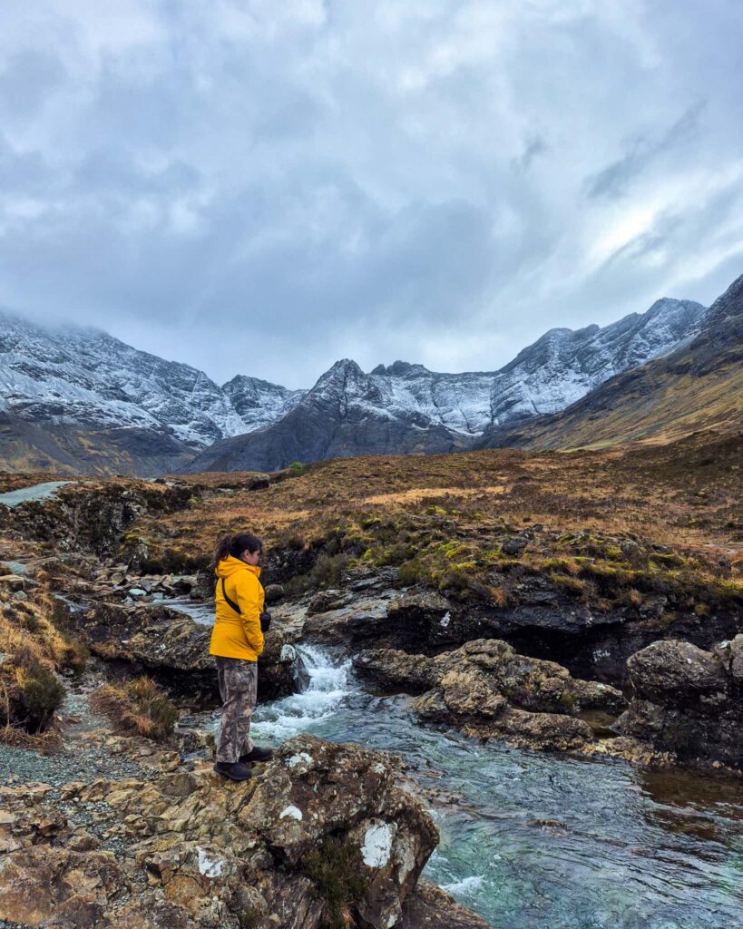 Alina looking down at the Fairy Pools in Isle of Skye, Scotland. She is wearing camo pants and a yellow rain jacket. There are snowy mountains in the background and a running stream.
