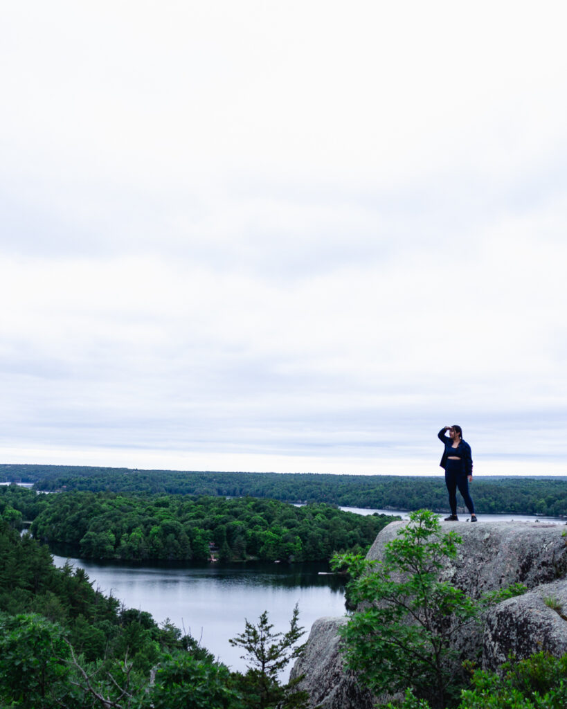 Alina, in an all blue athleisure outfit, overlooks the Rock Dunder summit. She stands on a cliff top, with green forests and a bay of water down below.