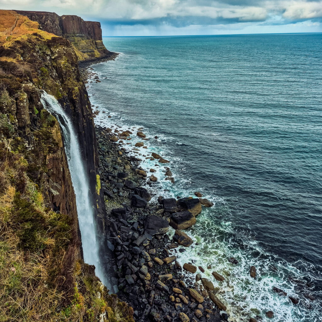 Sideview of Kilt Rock and Mealt Falls on the Isle of Skye in Scotland. A narrow waterfall pours out of a cliff face next to the Sea of the Hebrides.