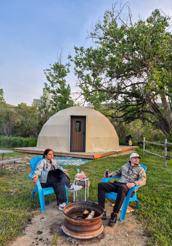 Alina and Justin sit in muskoka chairs in front of a geodome at Bridgeviews Drumheller