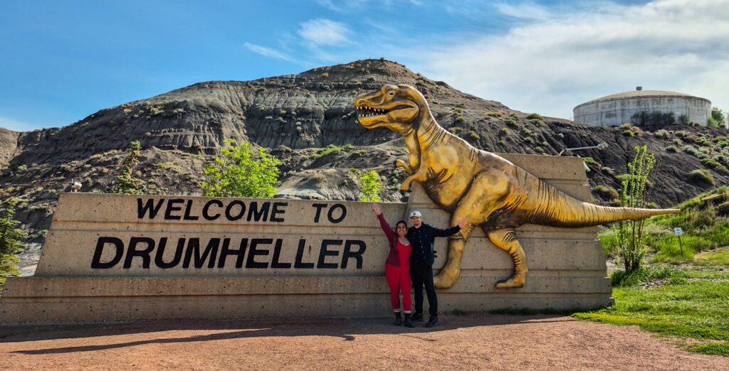 Alina and Justin standing with their arms up in front of the "Welcome to Drumheller" sign. There is a big gold t-rex on the sign.