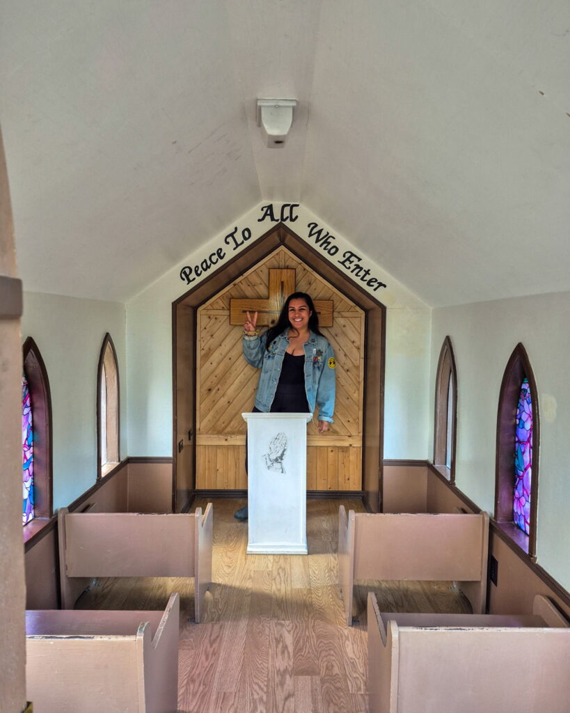 Alina standing at the pulpit inside Drumheller's Little Church. Inside is white with some small pews and purple stained glass in the windows. Behind Alina is a cross and the wall says "Peace To All Who Enter".