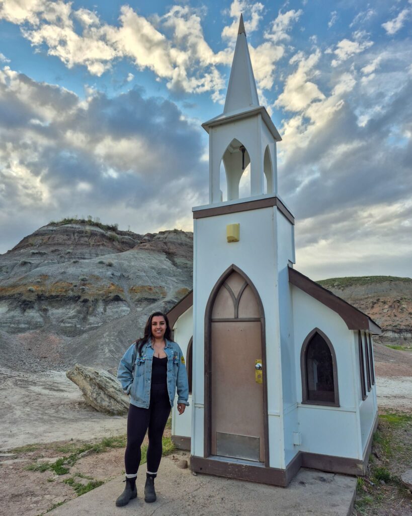 Alina standing outside the Drumheller Little Church. It is a tiny white church with a steeple. The roof and door are brown.