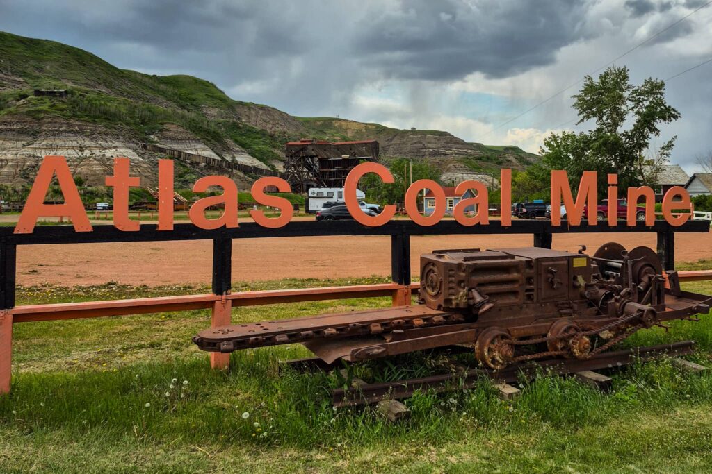 Orange "Atlas Coal Mine" Welcome Sign with some old machinery in front and wooden tipple in background.