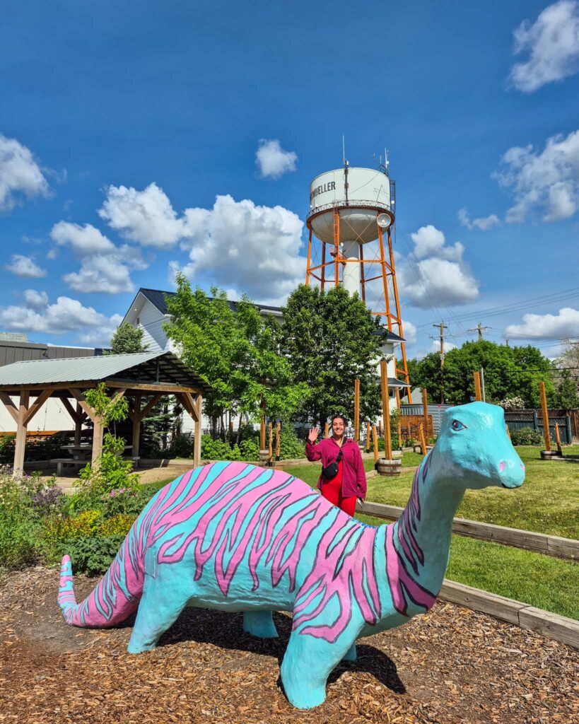 Alina standing behind a statue of a mint green brontosaurus with pink stripes. The Drumheller water tower is in the background.