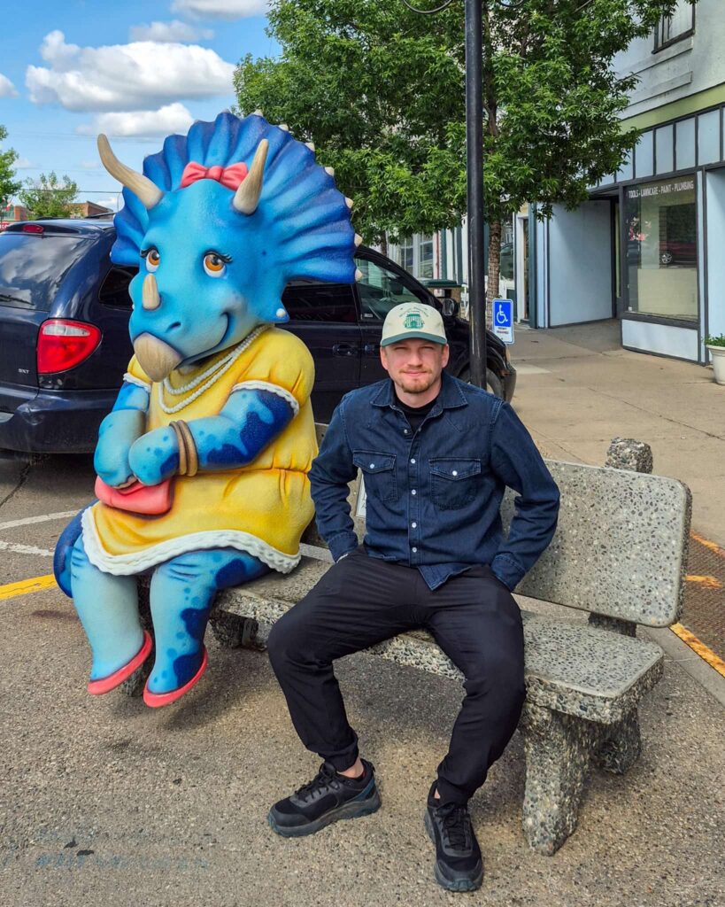 Justin sitting on a stone bench next to a triceratops statue also sitting on the bench. The triceratops status is blue and is wearing a yellow dress with pink accessories.
