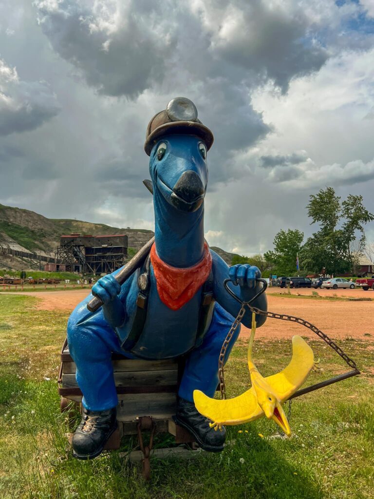 A blue dinosaur statue dressed as a miner complete with a denim outfit, a mining hat, and a pickaxe. The dinosaur holds a yellow pterodactyl in his other hand.