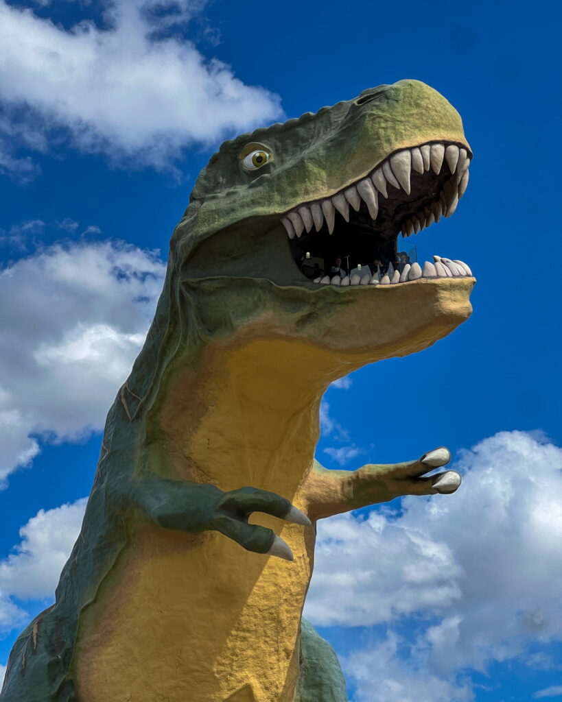 Tyra, the World's Largest Dinosaur. She is a giant green T-Rex statue with a yellow belly. Inside her jaws you can see people looking over the town of Drumheller.
