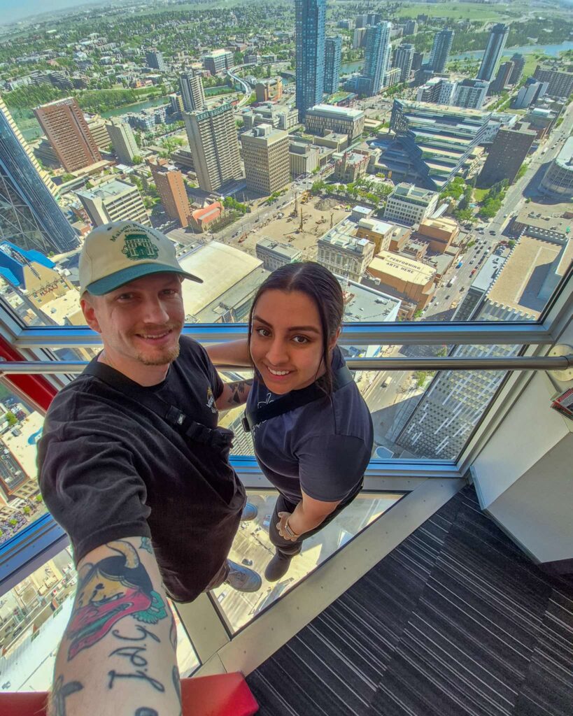 Alina and Justin taking a selfie inside the Calgary Tower. They are standing on a glass floor and you can see Calgary's downtown buildings below.