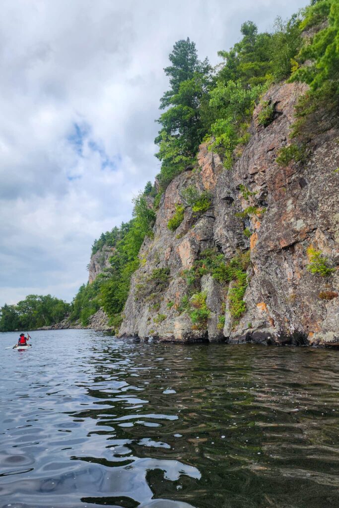 Paddling alongside Mazinaw Rock. There is a person sitting on a paddle board in the water ahead.