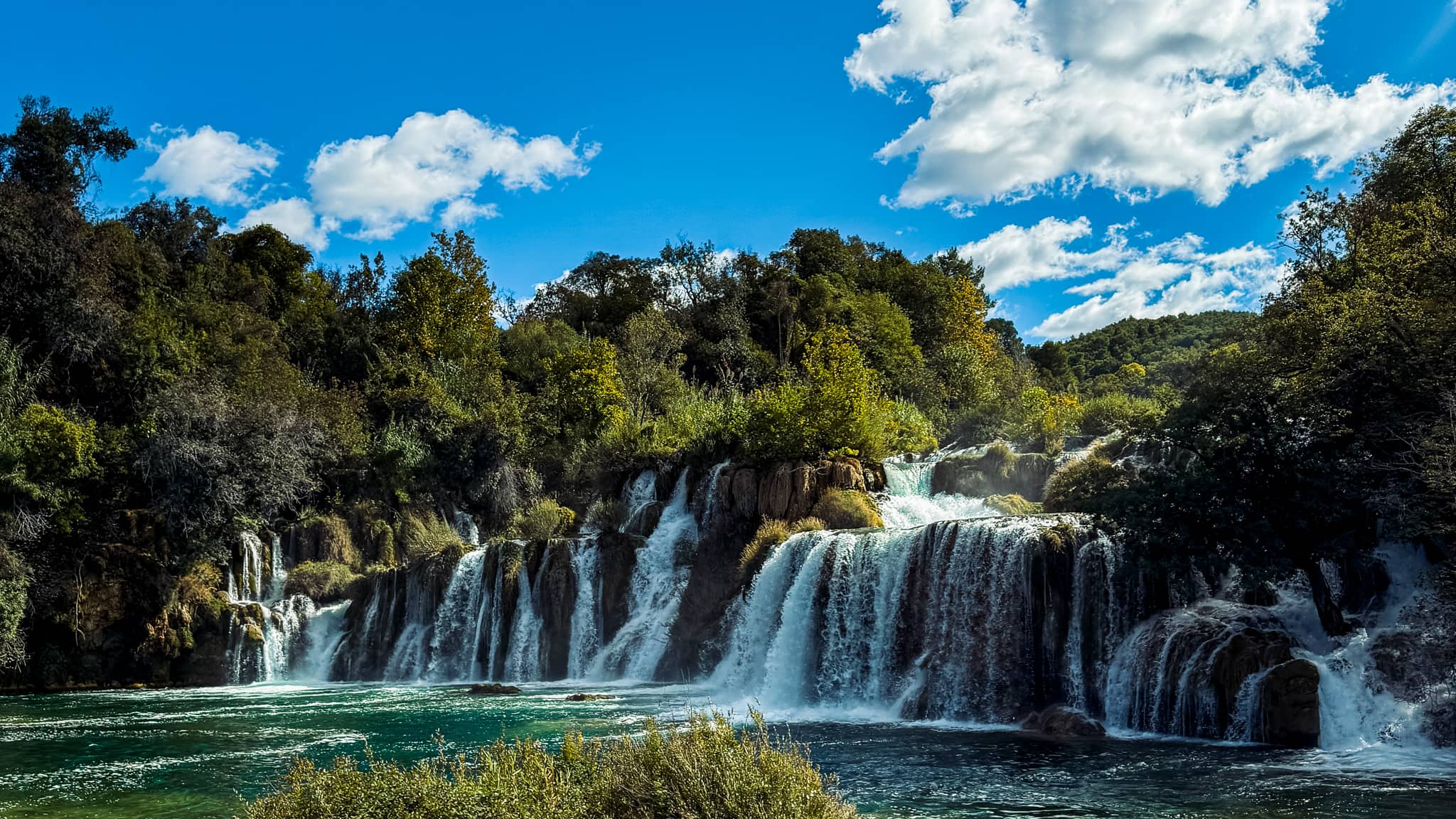 Skradinski Buk Waterfall. This is a multi-tiered waterfall surrounding by lush green trees, pouring into emerald coloured water.