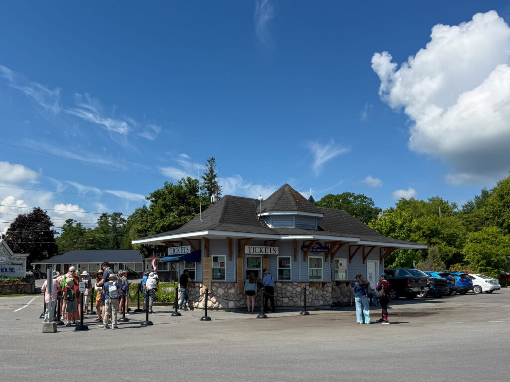 Rockport Cruises' Ticket Office. It is a small blue building with a black roof. A crowd of people is waiting in front to pick up their tickets.